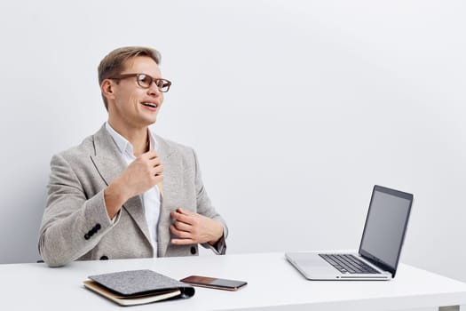 smiling young caucasian man wearing glasses and gray blazer sitting at white desk with laptop, phone, notebook, and remote control, looking aside while talking during video call about working remotely as digital nomad in modern white office. Concept: digital nomad