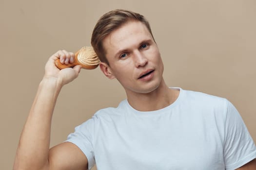 Relaxed young man with light brown hair and blue eyes, dressed in a simple white t-shirt, posing against a beige backdrop, holding a wooden brush near his ear. Studio shot emphasizing grooming, style, personal care, male lifestyle, natural pose, beauty care.