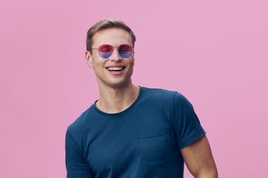 Smiling young man wearing colorful sunglasses and blue t-shirt looking away with cheerful expression against pink background. Casual lifestyle portrait showing positive mood and summer vibe. Concept: joy of summer.