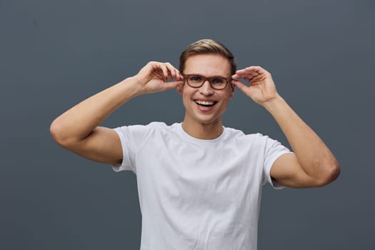 Smiling happy young caucasian man wearing glasses and white t-shirt looking at camera with joy isolated on dark gray background. Casual clothing and positive expression convey confidence and comfort. Concept: digital nomad lifestyle.