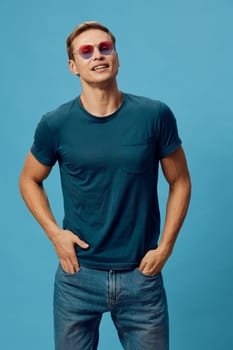 Cheerful man with sunglasses dressed in a casual blue t-shirt and denim jeans stands confidently against a vibrant blue backdrop. The relaxed pose and bright colors convey a fun, carefree summer fashion vibe, perfect for lifestyle and casual themes.