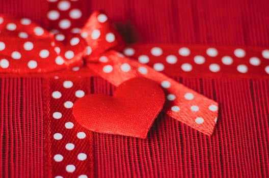 Close up of a red tiny heart and gift ribbon with white polka dots tied into a bow, placed on a textured red and pink striped background.