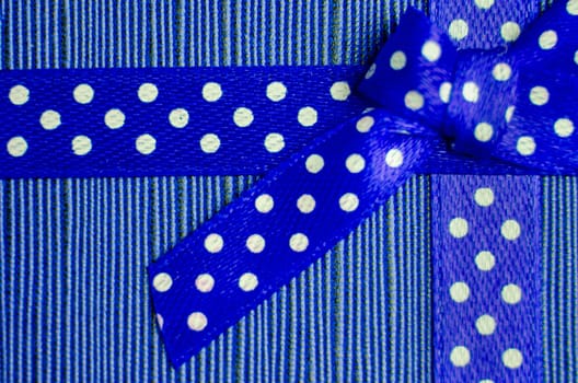 Close up of a blue gift ribbon with white polka dots tied into a bow, placed on a textured red and pink striped background.