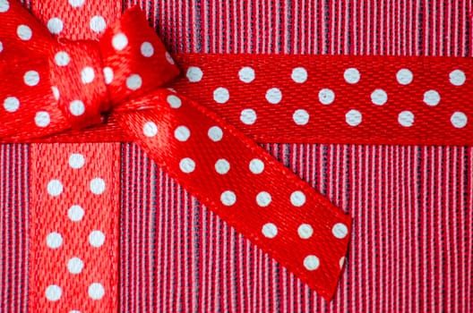 Close up of a red gift ribbon with white polka dots tied into a bow, placed on a textured red and pink striped background.