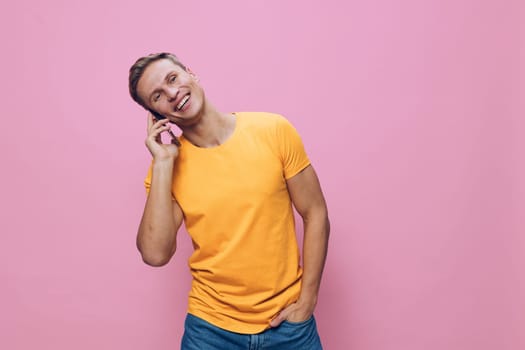 smiling man talking smartphone wearing orange t-shirt and jeans standing against pink background with hand in pocket enjoying digital nomad lifestyle and remote communication. Concept: digital nomad
