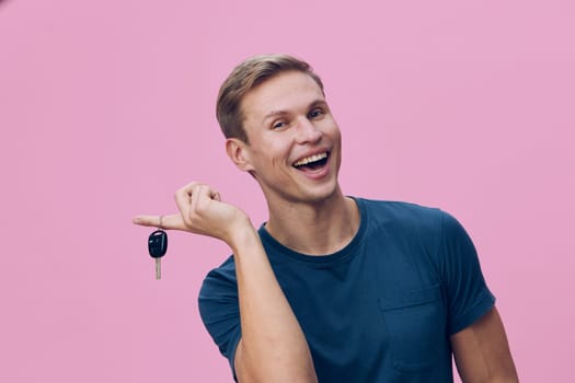 Cheerful man with short light brown hair, dressed in casual blue t-shirt, smiling broadly while holding car keys, standing against a plain pink background. Concept: happiness of new beginnings, personal success, celebration of achievement.