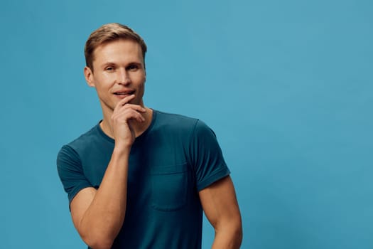 Joyful man with neat blond hair and casual blue t-shirt smiling thoughtfully while touching his chin, standing in front of a vibrant blue background; natural lighting, friendly mood, studio photography, positive energy.