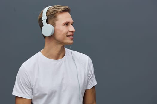 Smiling young caucasian man wearing white headphones and casual white t-shirt looking to the side against gray background. Relaxed expression, modern technology use, digital detox concept focusing on mental health and gadgets.