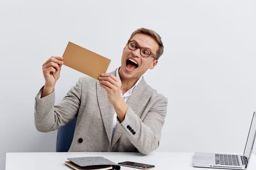 Excited young caucasian man holding envelope sitting at desk with laptop and planner, wearing eyeglasses and gray blazer, smiling joyfully. Concept of smart home automation, digital communication, and technology workflow.