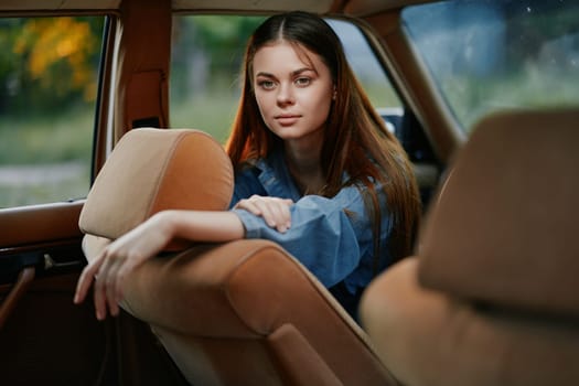 A young woman in a blue shirt sits in the back seat of a vintage car, gazing thoughtfully out the window. The warm colors of the interior contrast with the cool tones of the outdoor scenery, creating