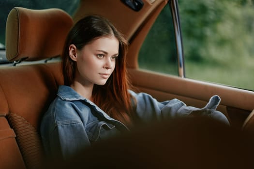 A thoughtful young woman dressed in denim relaxedly sits in a classic car, reflecting while enjoying the lush green scenery outside on a peaceful day.