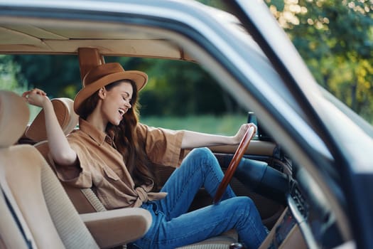 A cheerful young woman in a hat is joyfully sitting in a vintage car's interior, wearing a casual shirt and jeans, embodying freedom and adventure in a sunny green setting.