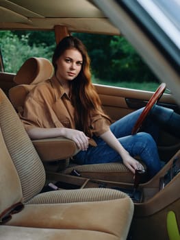 Stylish young woman with long hair sitting in a vintage car, dressed in casual clothing, conveying a sense of confidence and relaxation in a lush green setting.