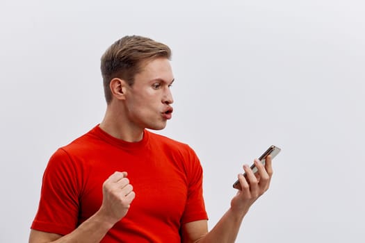 young man wearing red t-shirt use smartphone with excited facial expression fists clenched looking at screen close-up isolated light background digital detox and mental health gadgets concept