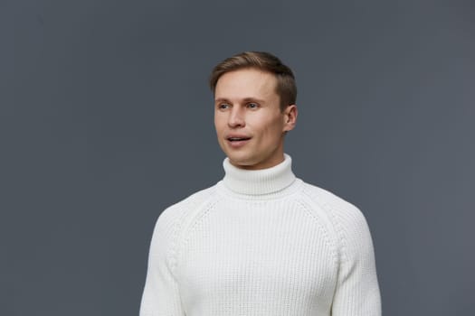 Caucasian man wearing white turtleneck sweater looking aside with slight smile on neutral gray background. Man close-up portrait, casual style, short hair, thoughtful expression. Concept: mental health and gadgets.