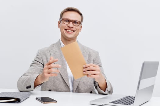 smiling caucasian man holding envelope sitting at table with laptop and mobile phone, engaged with digital communication, remote work and online correspondence, wearing glasses and casual jacket on white background. Concept: digital communication
