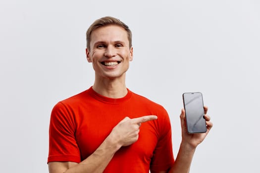 Cheerful young man with short hair in casual red t-shirt demonstrating smartphone with a happy expression on his face, standing against a plain neutral background. Concept: technology, communication, digital device