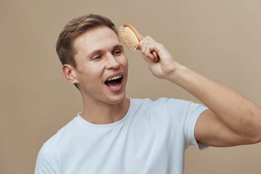 Happy young man with short brown hair brushing his hair with a wooden hairbrush, wearing a light blue t-shirt, posing against a neutral background, showcasing personal grooming, relaxed atmosphere, lifestyle portrait.