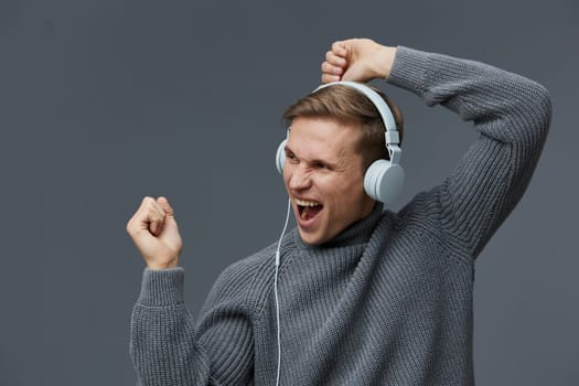 Happy man in casual gray sweater enjoying music with headphones, showing excitement and energy. Isolated gray backdrop emphasizes his joyful mood and relaxed style, lifestyle, celebrating feelings.