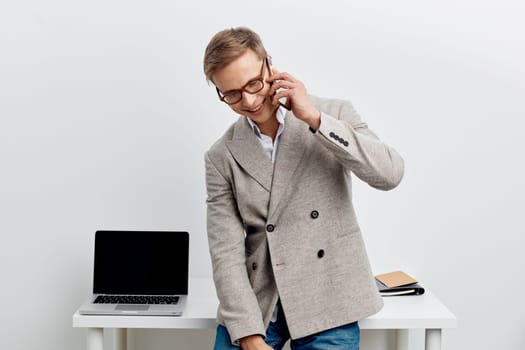 Friendly young man in smart casual clothing engaging in phone conversation at workplace, standing near desk with open laptop and notepad, cheerful expression, modern office environment with minimalist white background, concept: professional communication.