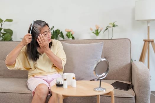 A mature woman applies skincare while combing her hair, highlighting her beauty routine in a cozy living room setting.