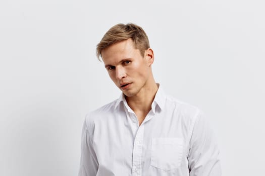 Stylish young man with short hair and fair skin, dressed in a white shirt, posing with a slight tilt, looking at the camera with a serious expression. Background is plain and neutral, emphasizing his confident mood and modern appearance. Concept: professionalism and style.