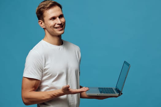 Young cheerful man in a white t-shirt demonstrating a laptop against a blue background, conveying digital communication, online work, modern lifestyle, technology use, casual style, studio portrait.