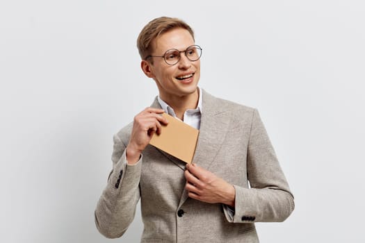 Happy young man with glasses in a gray blazer holding a notepad, smiling and looking away, casual yet professional appearance, indoor studio portrait with a plain light background, conveying confidence and optimism. Concept: professional success