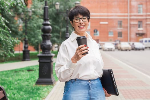 Senior woman enjoying coffee during walk with laptop. Urban lifestyle, freelance work and active aging.