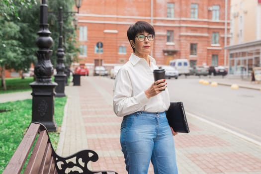 Mature woman with coffee and laptop standing outdoors. Freelance work, career focus and independent lifestyle.