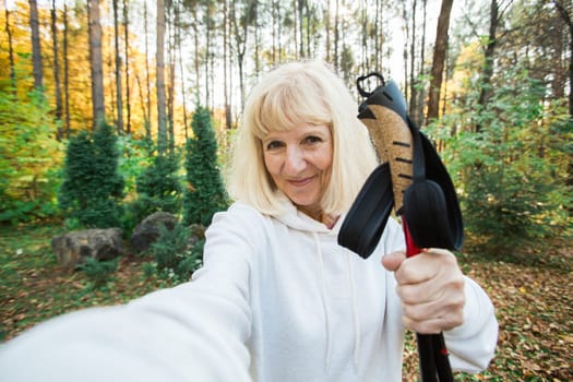 Senior woman taking a cheerful selfie during a nordic walk and take selfie on smartphone. Concept of active lifestyle, adventure, fitness, freedom and joy in the golden autumn forest.