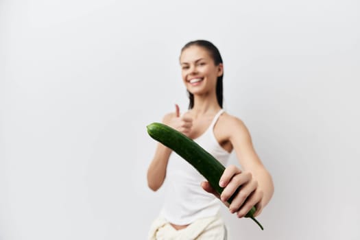 Smiling woman in a simple white tank top holds a cucumber while giving a thumbs up The bright, neutral background emphasizes the healthy lifestyle theme, perfect for wellness and nutrition concepts