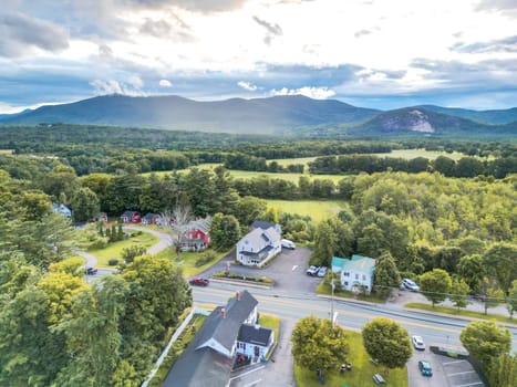 A picturesque small town in a valley, featuring vibrant greenery and towering mountains, with a clear sky and a peaceful road lined with parked cars, creating a serene atmosphere.