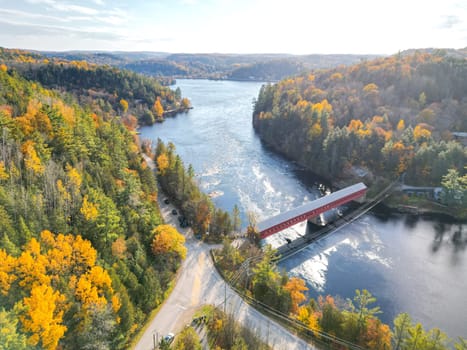 The image shows a small red bridge over a river in a beautiful autumn forest. The trees are in full foliage and the leaves are turning yellow, orange, and red. The water is calm and crystal clear.