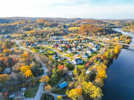 A beautiful small town filled with charming houses, vibrant trees, and a serene river flowing through it. The sky is a brilliant blue adorned with fluffy white clouds, and the sun shines brightly.