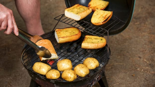 A close-up shot of a charcoal grill with a grate full of potatoes and garlic bread. The potatoes are various sizes and the garlic bread is made with thick slices of bread.