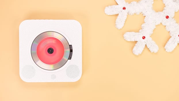 A white portable CD player is positioned on a beige background next to a decorative white snowflake made of tinsel, with a red light shining from inside the CD player.