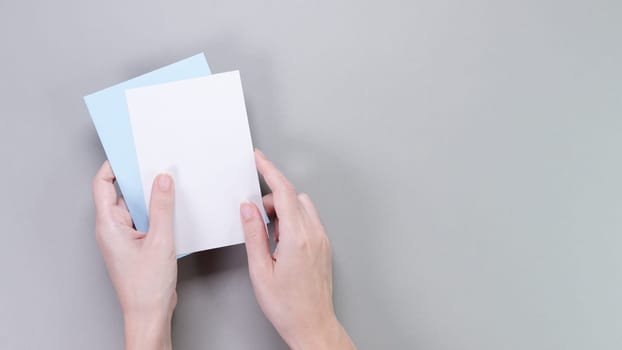 The image showcases a pair of slender female hands elegantly holding two blank note cards slightly apart. The cards are set against a solid gray background, creating a clean and modern aesthetic.