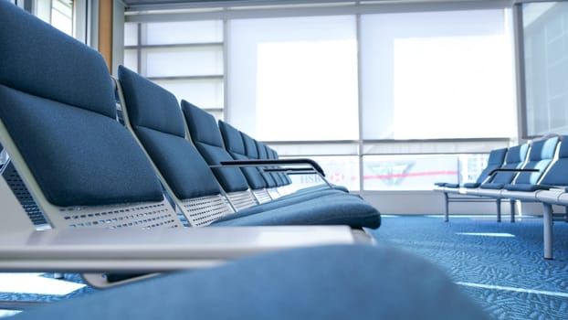 This is a photo of a row of empty blue airport seats with a large glass window in the background. The photo is taken from the perspective of a passenger sitting in one of the seats.