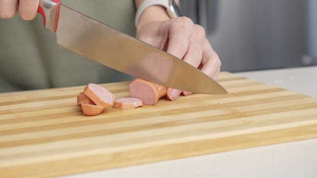 Close-up of a woman cutting sausage on a wooden cutting board in a kitchen setting with different ingredients