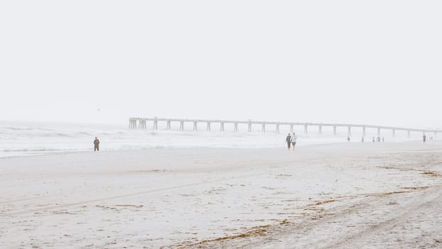 People walking on a beach with a pier in the distance on a foggy day. Jacksonville, Florida USA 03.15.2024