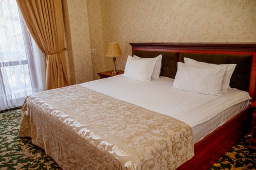 Hotel room with golden yellow blanket on dark red bed and green carpet on the floor. Room in golden beige tones with white bed linen. on the bedside table table lamp. door to the balcony with curtain