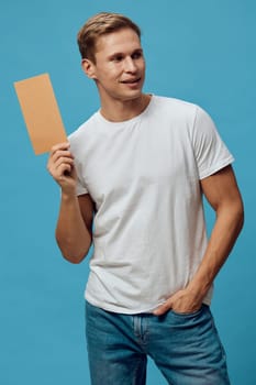 smiling man holding cardboard piece wearing casual white t-shirt and jeans standing on blue background looking aside with relaxed posture modern digital nomad lifestyle concept related to communication and creativity