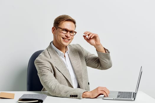 A cheerful young man in glasses, dressed in a light blazer and white shirt, sitting at a desk with a laptop, smiling warmly at the camera. Modern office setting, positive mood, professional atmosphere.