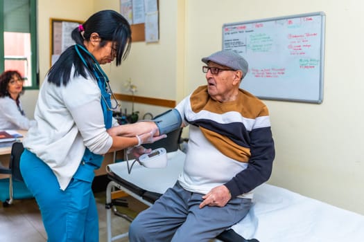 Female nurse checking blood pressure of elderly man sitting on examination table in medical clinic