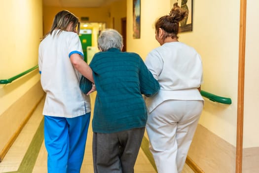 Two nurses are assisting an elderly woman as they walk down the hallway of a nursing home, providing support and care