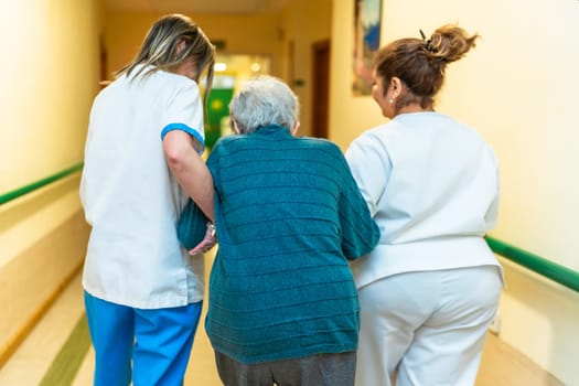 Two nurses are assisting an elderly woman as they walk down the hallway of a nursing home, providing support and care
