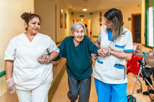 Two nurses assisting an elderly woman walking down the corridor of a nursing home, providing support and care