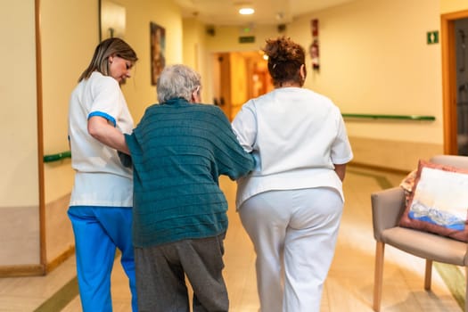 Two nurses are assisting an elderly woman as they walk down the hallway of a nursing home, providing support and care