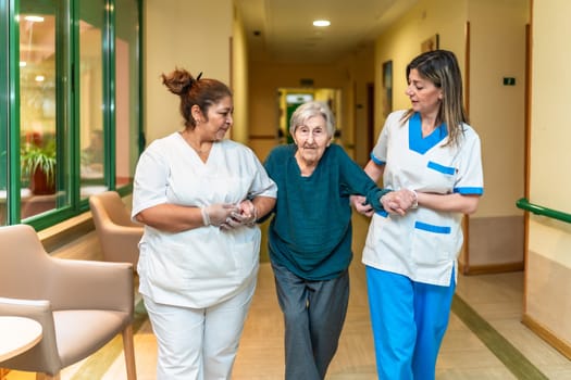 Two nurses assisting an elderly woman walking down the corridor of a nursing home, providing support and care